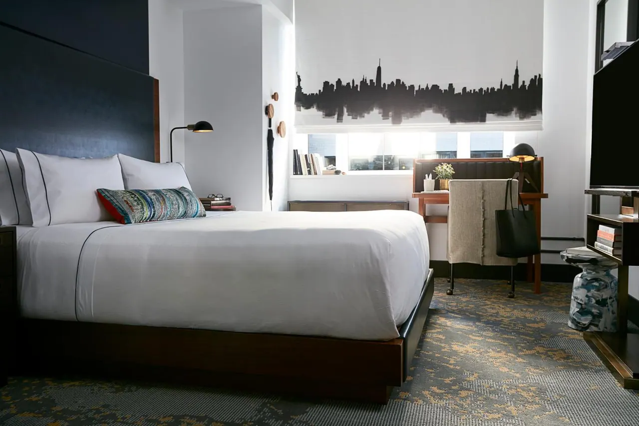 A king bed with white linens and a patterned cushion sits beside a desk and television, with a skyline-printed window blind behind it in a hotel bedroom.