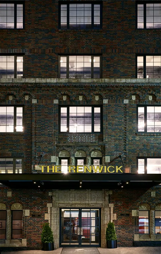 Night view of the brick facade of The Renwick hotel, with illuminated yellow lettering above a glass entrance door flanked by two potted shrubs.