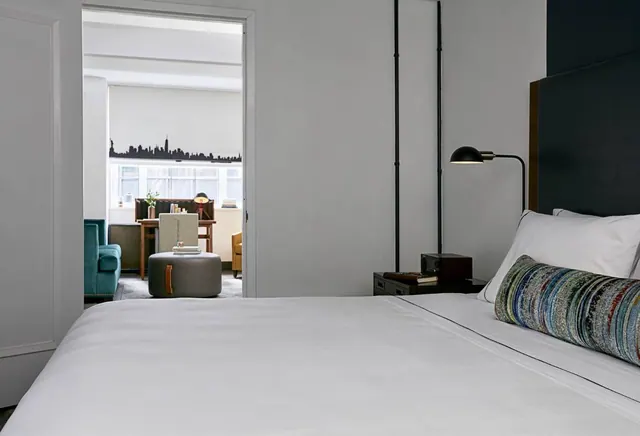 A made king bed with white linens and a striped bolster sits beside a nightstand with a lamp, with a sitting area and desk visible through the open doorway.