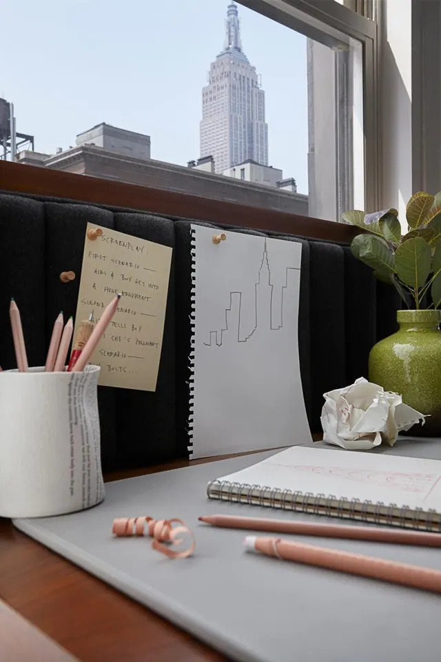 A desk by a window holds pencils, a notepad, a crumpled paper and a green vase, with the Empire State Building visible through the glass.