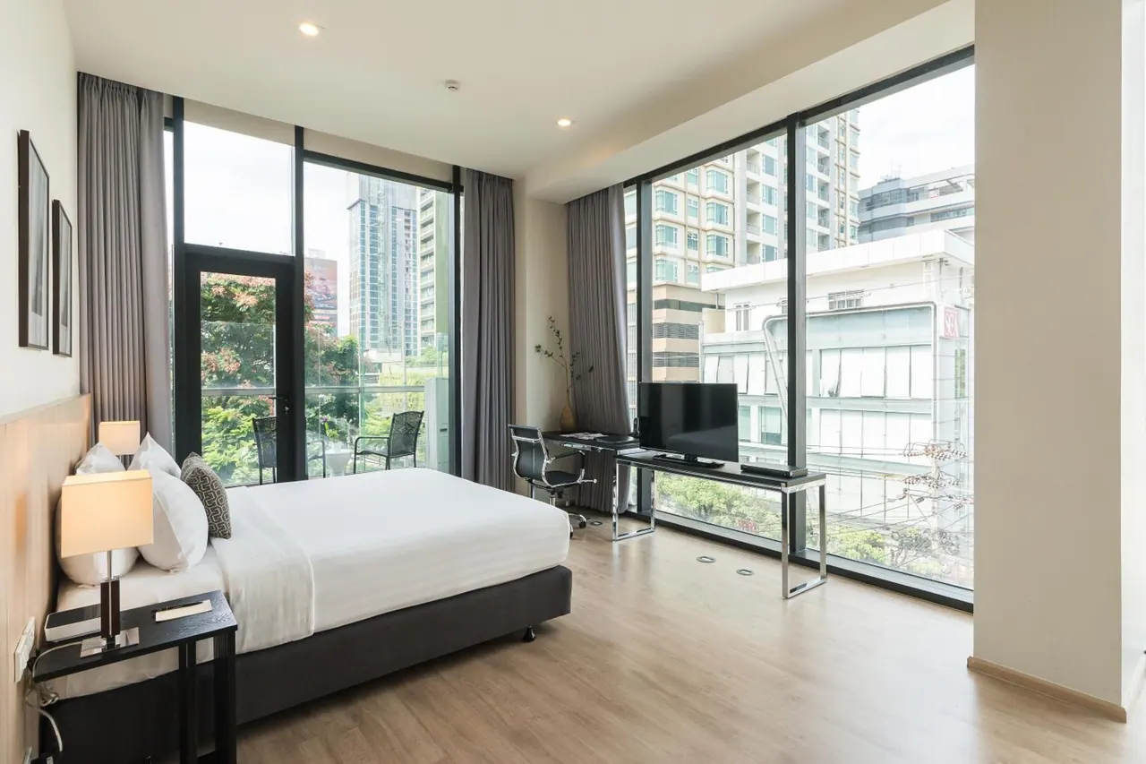 A bedroom with a king bed, a work desk with a television, and floor-to-ceiling windows and a glass door opening onto a balcony with city towers beyond.
