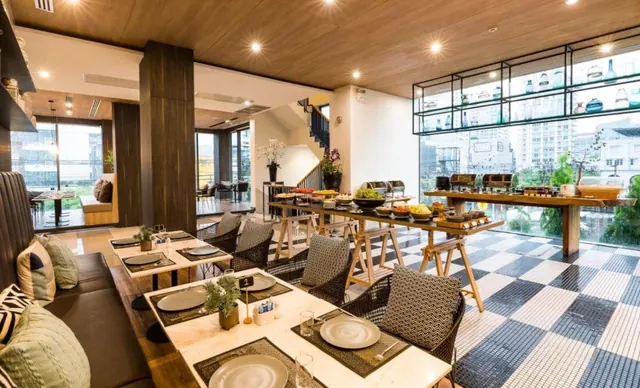 A restaurant dining room with set tables, a buffet counter on wooden trestles, a checkered floor and floor-to-ceiling windows overlooking high-rise buildings.