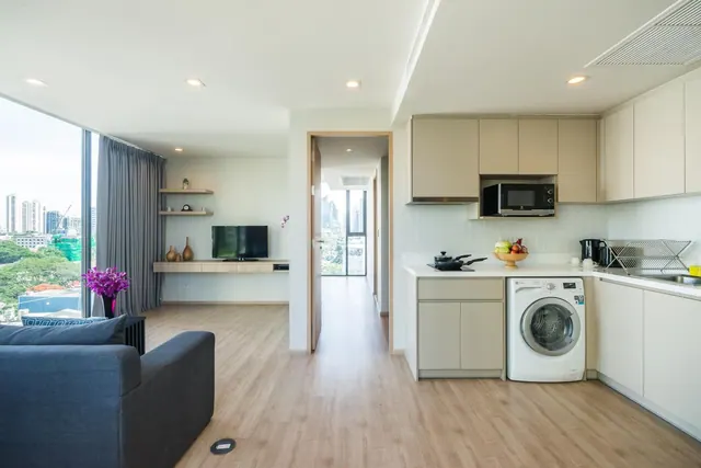 A living area with a grey sofa, wall-mounted television and shelving, an open kitchen with a washing machine, and city buildings visible through a large window.