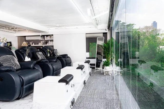 A pedicure area with black leather recliners and white foot basins beside a floor-to-ceiling glass wall that looks out onto trees.