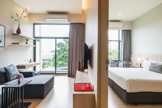 A hotel room living area with a grey sofa, a wall-mounted television and floor-to-ceiling windows looking onto trees, with a bed and desk visible through the doorway.