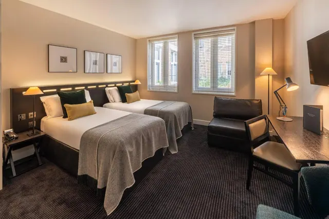 A twin bedroom with two beds, a leather armchair, a wooden desk with a desk lamp, and a wall-mounted television, with windows looking onto a brick building.
