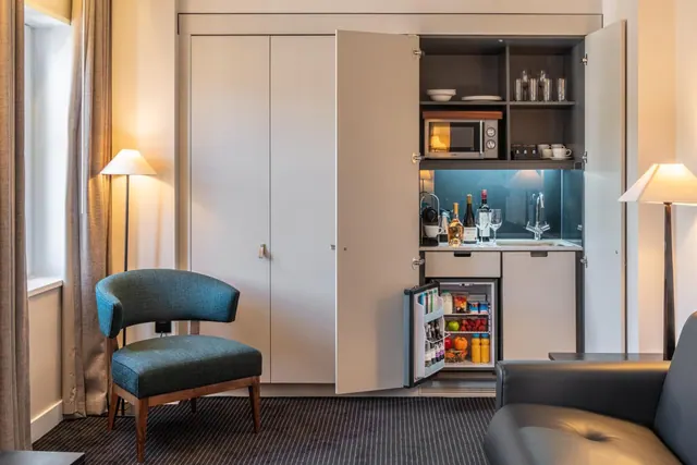 A hotel room living area with a teal armchair, a grey leather sofa, and an open built-in kitchenette holding a microwave, coffee machine, sink and minibar fridge.