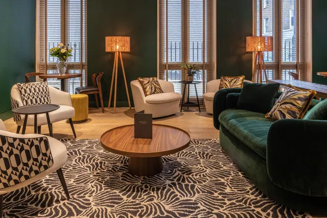 A lounge seating area with a green velvet sofa, patterned armchairs, a round wooden coffee table with a menu card on it, floor lamps and tall windows with blinds.