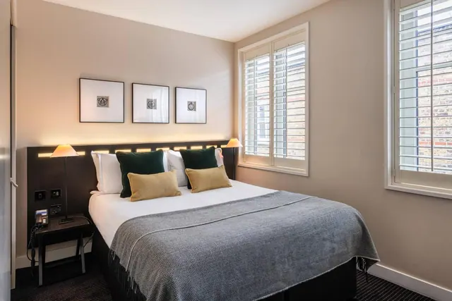 A bedroom with a double bed dressed in white linen, a grey throw and green and gold cushions, with shuttered windows looking onto a brick wall.
