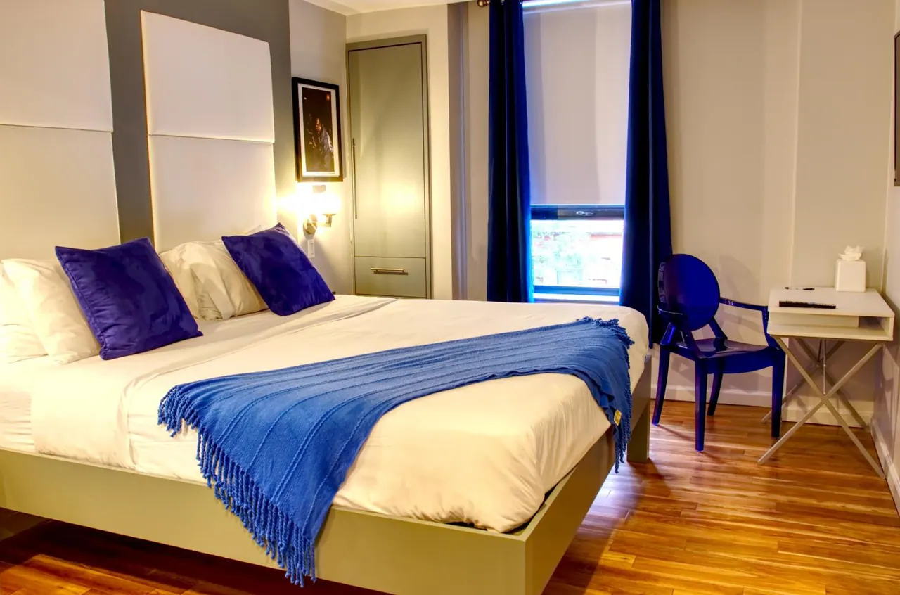 A bedroom with a made king bed in white linen and blue cushions and throw, blue curtains by the window, a blue chair and a small writing desk on a wooden floor.