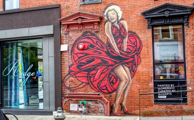 Exterior of the Ridge Hotel on a red brick facade with a large red mural of a woman in a flowing dress, a glass entrance door with the hotel name, and a sidewalk in front.