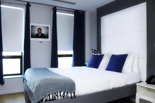 A bedroom with a double bed dressed in white linen and blue cushions, with a grey patterned throw, blinds and dark curtains on the windows, and a framed portrait on the wall.