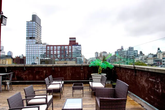 A rooftop terrace with wicker armchairs, low glass-top tables, a potted palm and a view of city buildings beyond a glass railing.