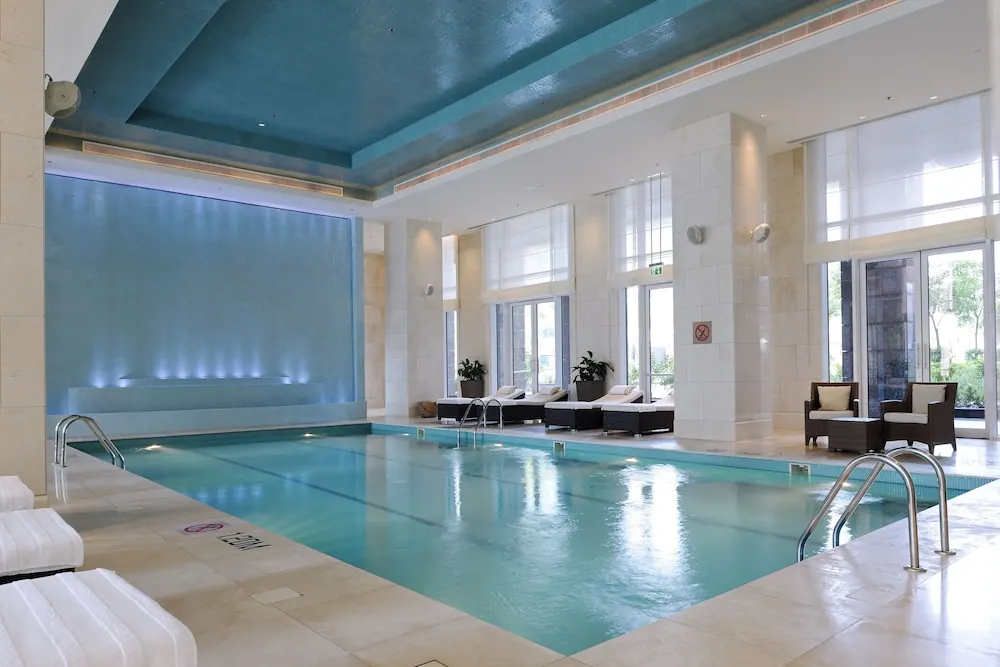 An indoor lap pool with lane markings, a backlit blue wall, white loungers and armchairs beside tall windows.