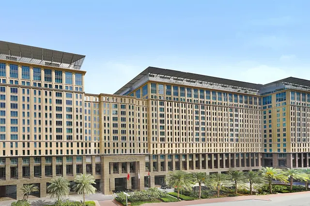 The exterior of a large sand-coloured hotel building with palm trees along the front under a clear blue sky.