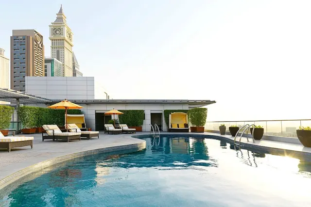 A rooftop swimming pool with a curved edge, sun loungers, yellow parasols and cabanas, with city towers visible beyond the railing.