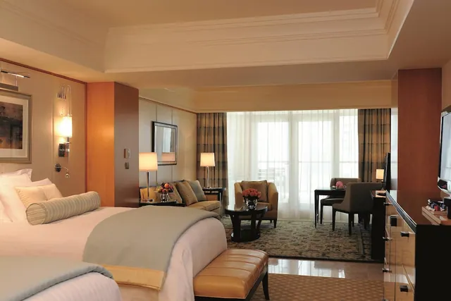 A hotel bedroom with a large bed, a sofa and armchairs by sheer-curtained windows, a desk, and a wall-mounted television.