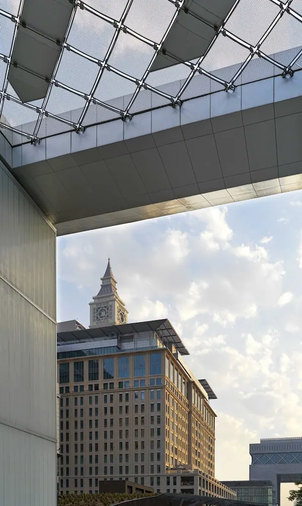 Looking up at a metal and mesh canopy with a clock tower and office buildings visible beyond under a partly cloudy sky.