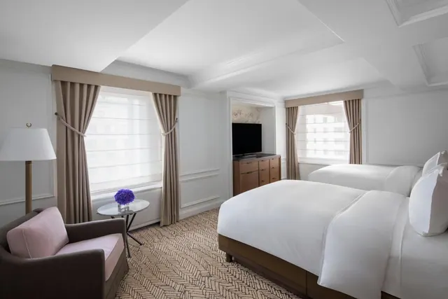 A hotel bedroom with two beds, a pink-cushioned armchair, a side table with purple flowers, a floor lamp, and a TV on a wooden cabinet.