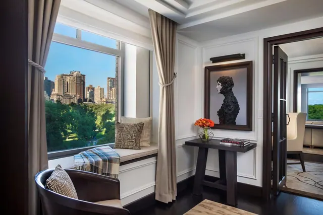 A window seat with cushions and a plaid throw overlooks green trees and city high-rises, beside a console table with a framed sculpture and an open doorway to another room.