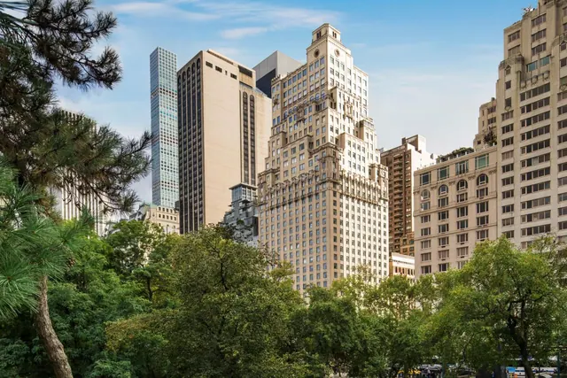 The Essex House tower and neighbouring skyscrapers rise behind trees in the foreground under a partly cloudy sky.