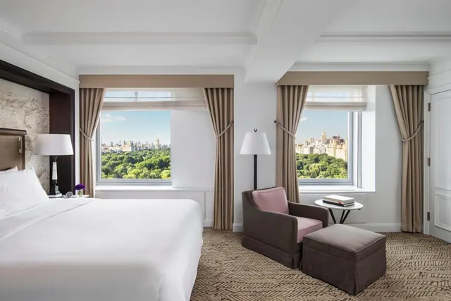 A hotel bedroom with a king bed, a pink armchair and ottoman, and two windows overlooking green trees and city towers.