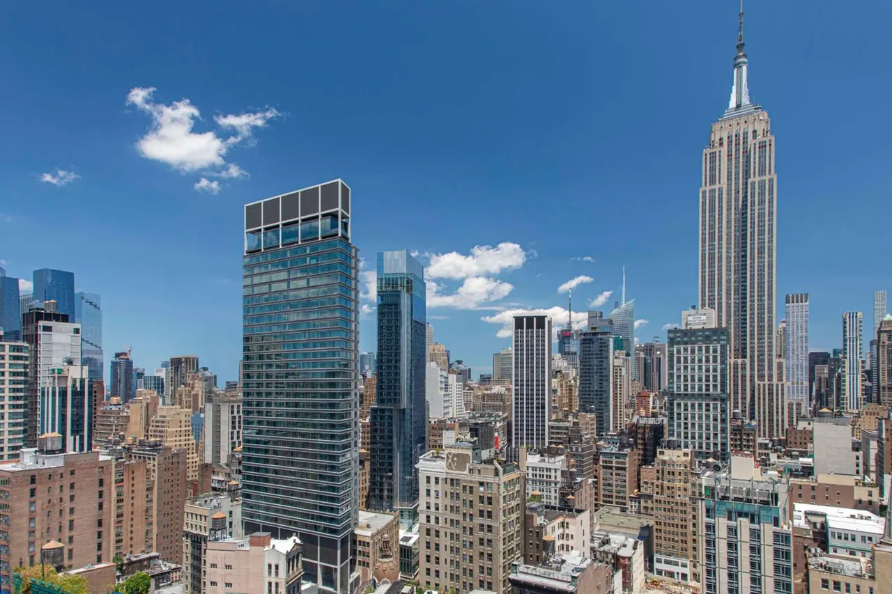 Elevated view over dense Manhattan buildings toward the Empire State Building under a blue sky with scattered clouds.