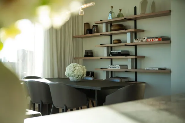 A dining area with a dark wooden table, grey upholstered chairs, a white flower arrangement and wall shelves holding books and decorative objects, with city buildings visible through the window.