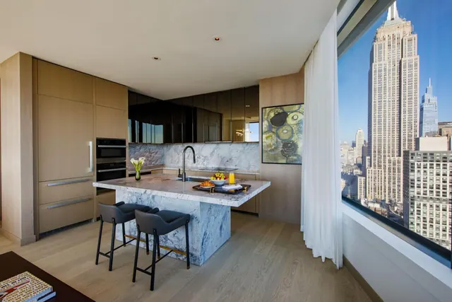 A kitchen with a marble-topped island and two bar stools sits beside a floor-to-ceiling window showing the Empire State Building.