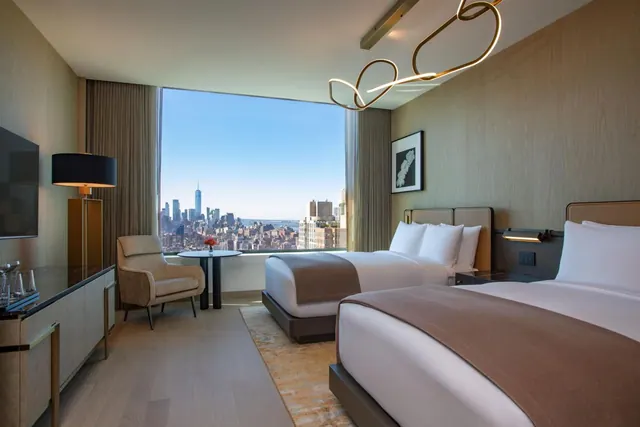 A hotel room with two made beds, an armchair and round table, and a large window showing the Manhattan skyline with One World Trade Center.