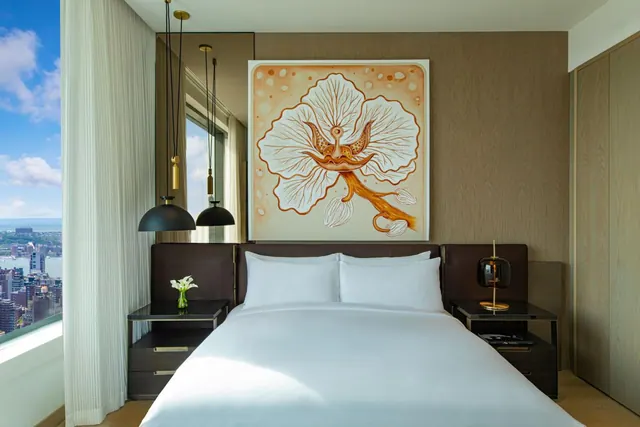 A bedroom with a made king bed, a large framed painting above the headboard, pendant lights, and a window showing a city skyline and water.