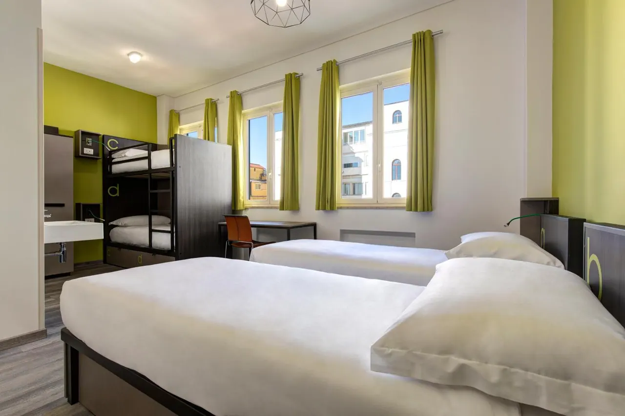 A bedroom with two single beds in the foreground, a bunk bed on the left, a desk, and green curtained windows looking onto a neighbouring building.