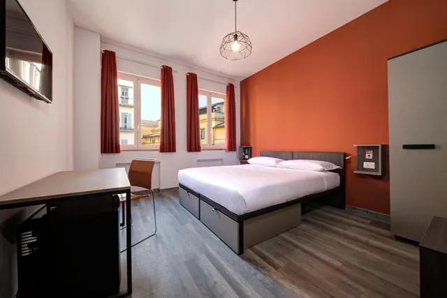 A double bed stands against an orange accent wall, with a desk, wall-mounted television and windows looking onto neighbouring buildings.