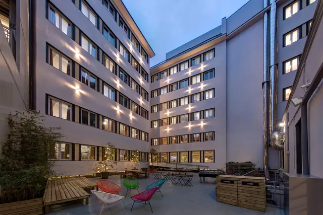 An enclosed courtyard at dusk with lit windows on the surrounding building, colourful chairs, small tables, a wooden bench and potted bamboo plants.