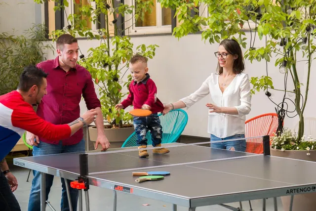 Four people play table tennis on an outdoor table in a courtyard with potted plants, string lights and colourful chairs.