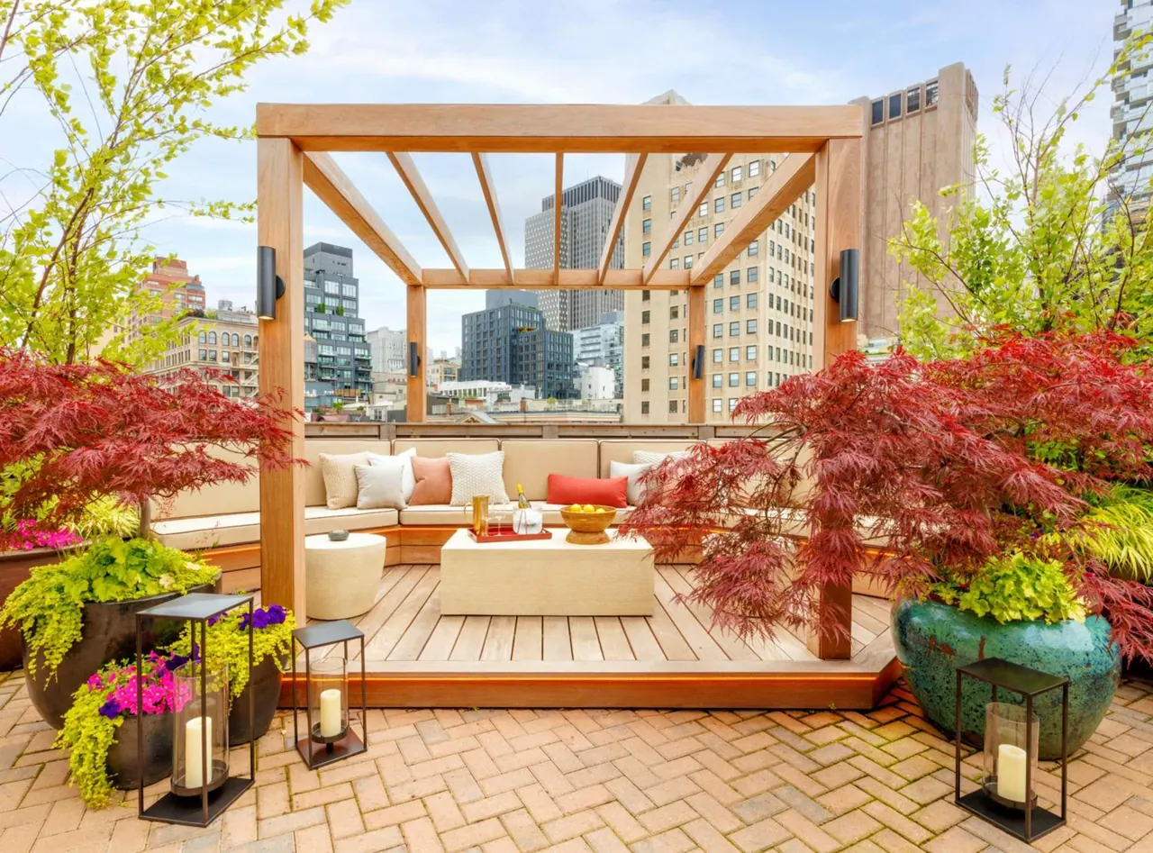 A rooftop terrace with a wooden pergola, a cushioned sectional sofa, a coffee table, potted Japanese maples and candle lanterns, with city high-rises visible behind.