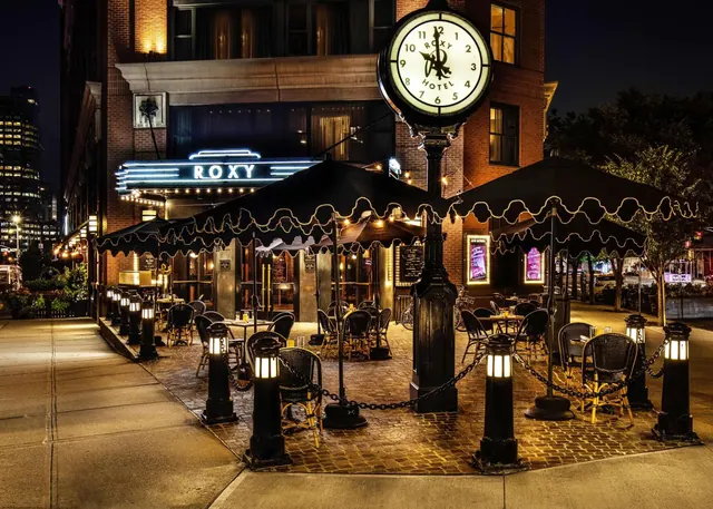 Illuminated sidewalk seating with black umbrellas, chained bollards and a lit street clock stands outside the Roxy Hotel building at night, with a neon sign on the facade.