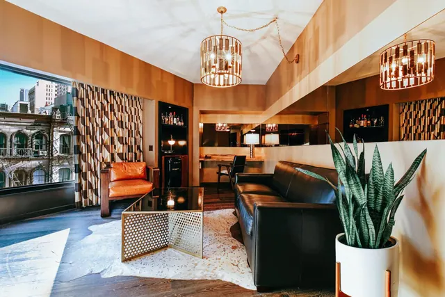 A living area with a black leather sofa, an orange armchair, a cowhide rug, two pendant chandeliers, and a large window showing city buildings.