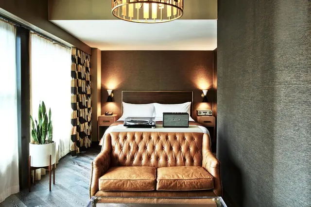 A bedroom with a double bed, a turntable and speaker on the bed, a tufted brown leather loveseat in the foreground, a potted snake plant and sheer curtains by the window.