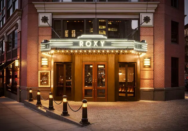 The front entrance of the Roxy hotel at dusk, with a lit neon marquee above a brick facade, wooden glass doors, and chained bollards on a paved plaza.