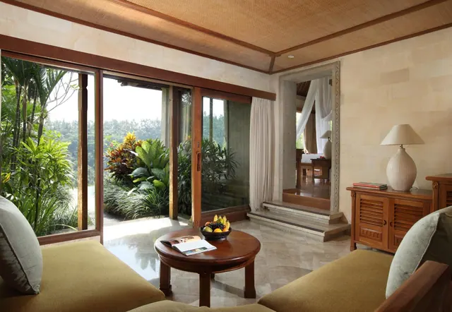 A living area with sliding glass doors open onto a tropical garden, with a round wooden coffee table, sofas, and a view through to a bedroom with a canopy bed.
