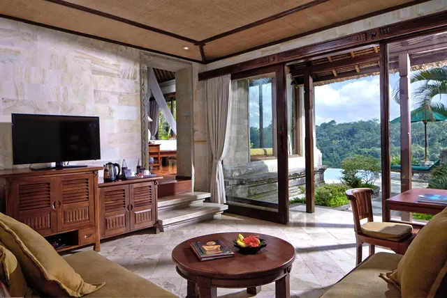 A living area with a flat-screen television on a wooden cabinet, a sofa, and a round coffee table with a fruit bowl, with glass doors opening onto a forested view and a pool.