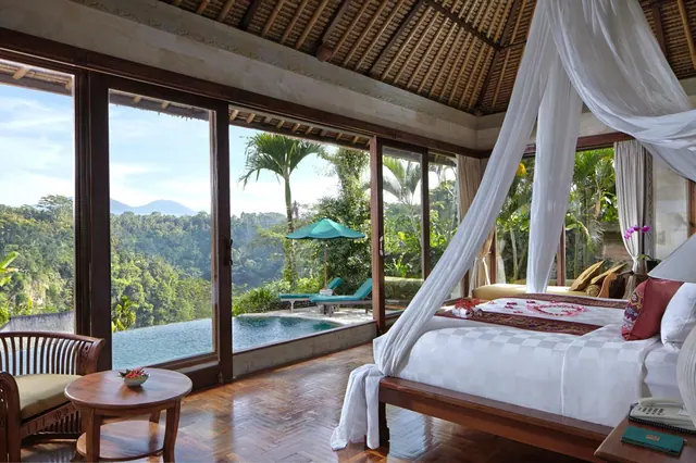 A bedroom with a canopy bed and floor-to-ceiling glass doors looking onto an infinity pool, sun loungers and a jungle valley with distant mountains.