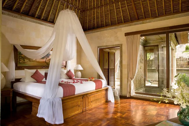 A bedroom with a canopied king bed dressed in white linen and red cushions, a thatched ceiling, a parquet wood floor, and glass doors looking onto a bamboo-fenced courtyard.