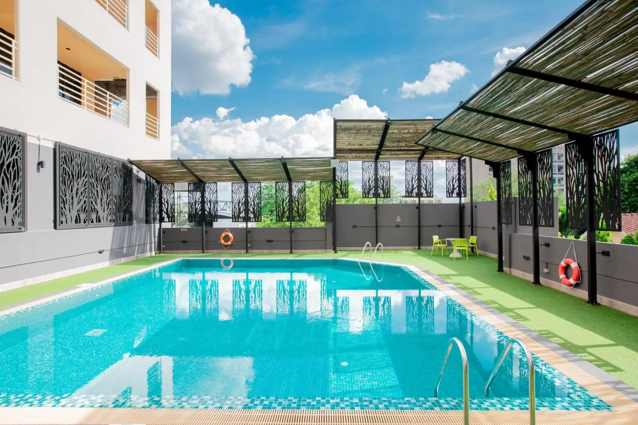 An outdoor swimming pool with turquoise water, green artificial turf around the edges, and a covered pergola-style shade structure along the right side.