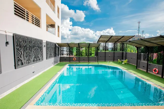 A rectangular outdoor swimming pool with turquoise tiles sits beside a building, with slatted pergola shelters, green artificial turf and a red lifebuoy on the wall.