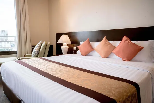A made king bed with striped white linen, a brown and gold runner and orange cushions sits beside a table lamp and an armchair, with a window showing city buildings at left.