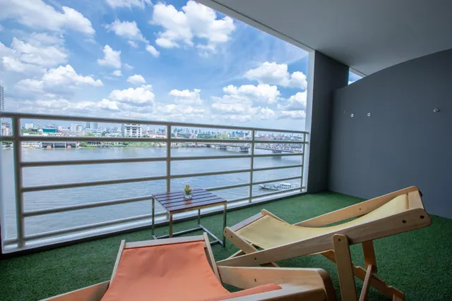 A covered balcony with two wooden deck chairs and a small side table overlooks a wide river and the city skyline across the water.
