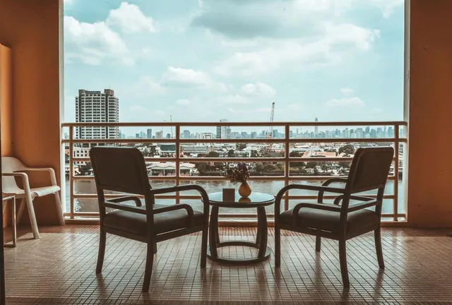 A tiled balcony with two armchairs and a small round table faces a river and city skyline.