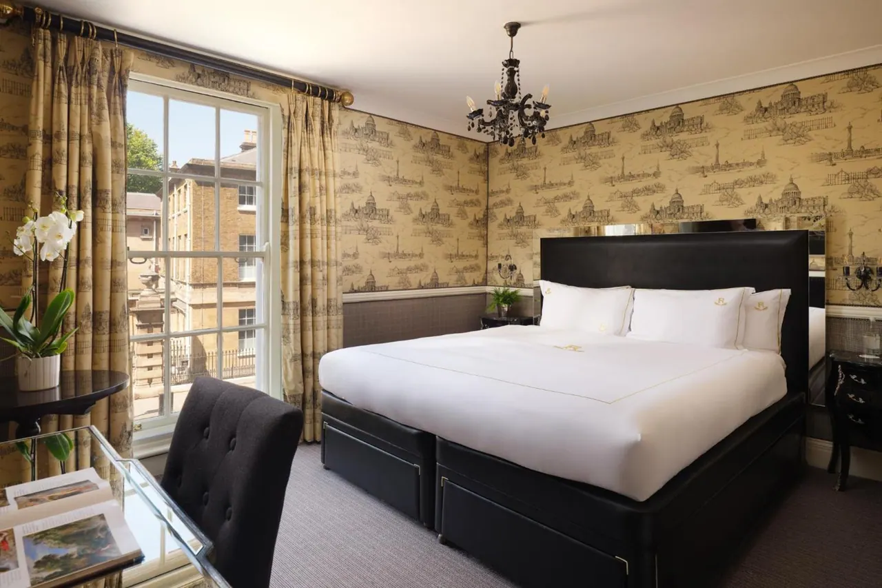 A bedroom with a made king bed under a black chandelier, a glass desk and chair by a sash window, and patterned wallpaper, with neighbouring buildings visible outside.